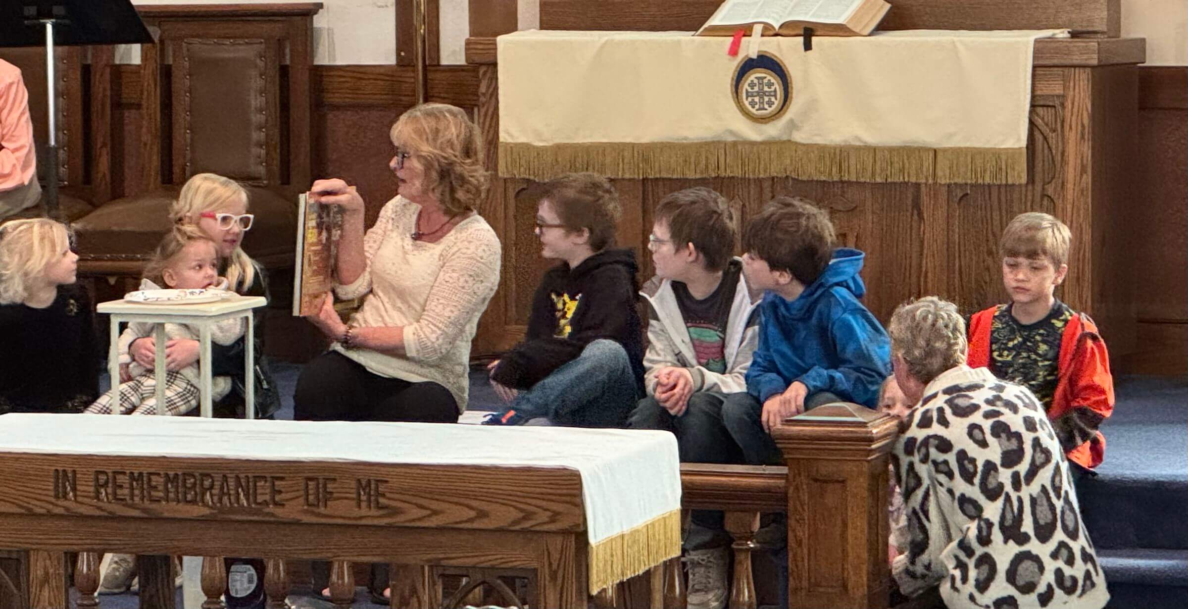A woman reads to a group of children seated on the floor in a church, near a wooden table with "IN REMEMBRANCE OF ME" inscribed on the front.