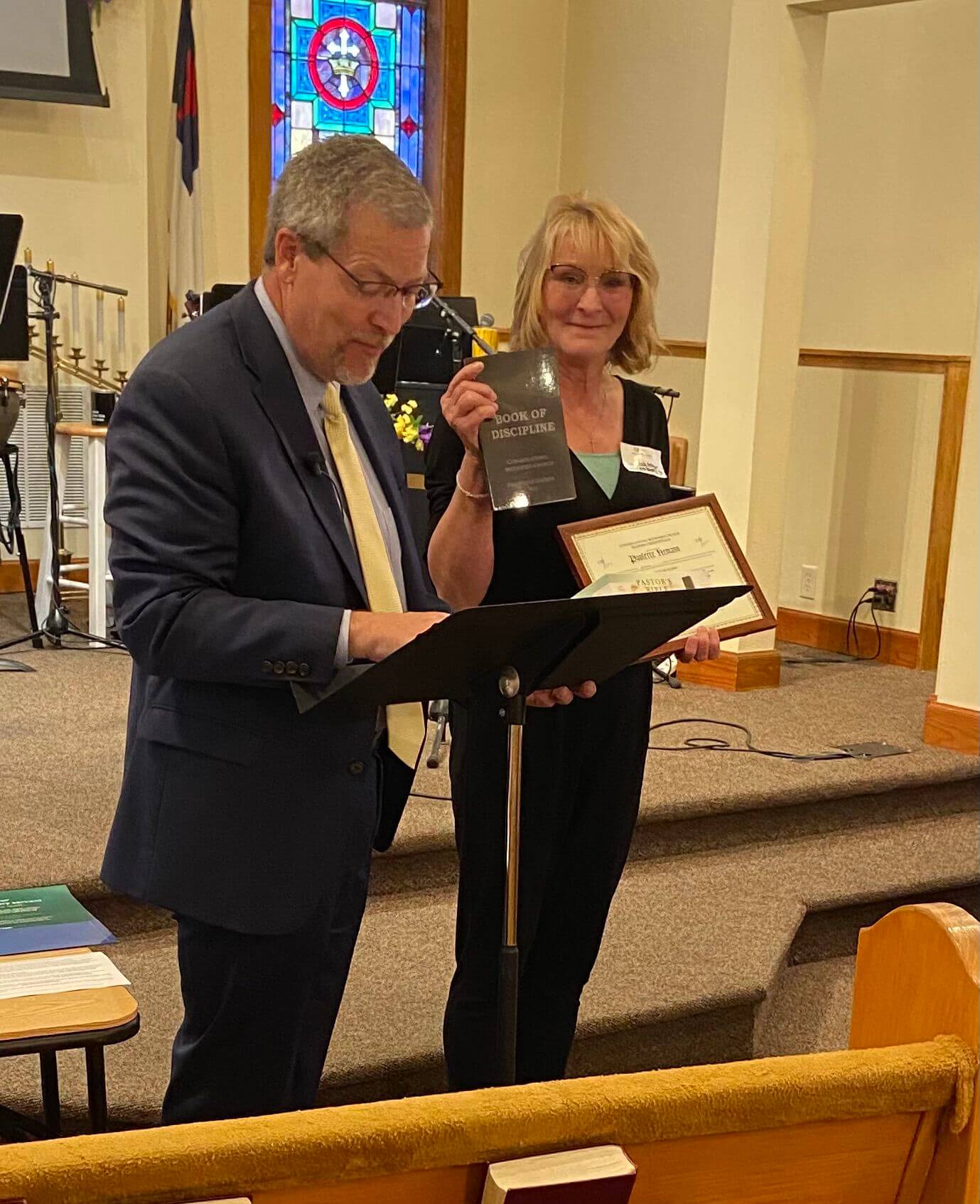 A man stands at a podium reading while a woman beside him smiles, holding a certificate and a book titled "Book of Discipline" in a church setting.
