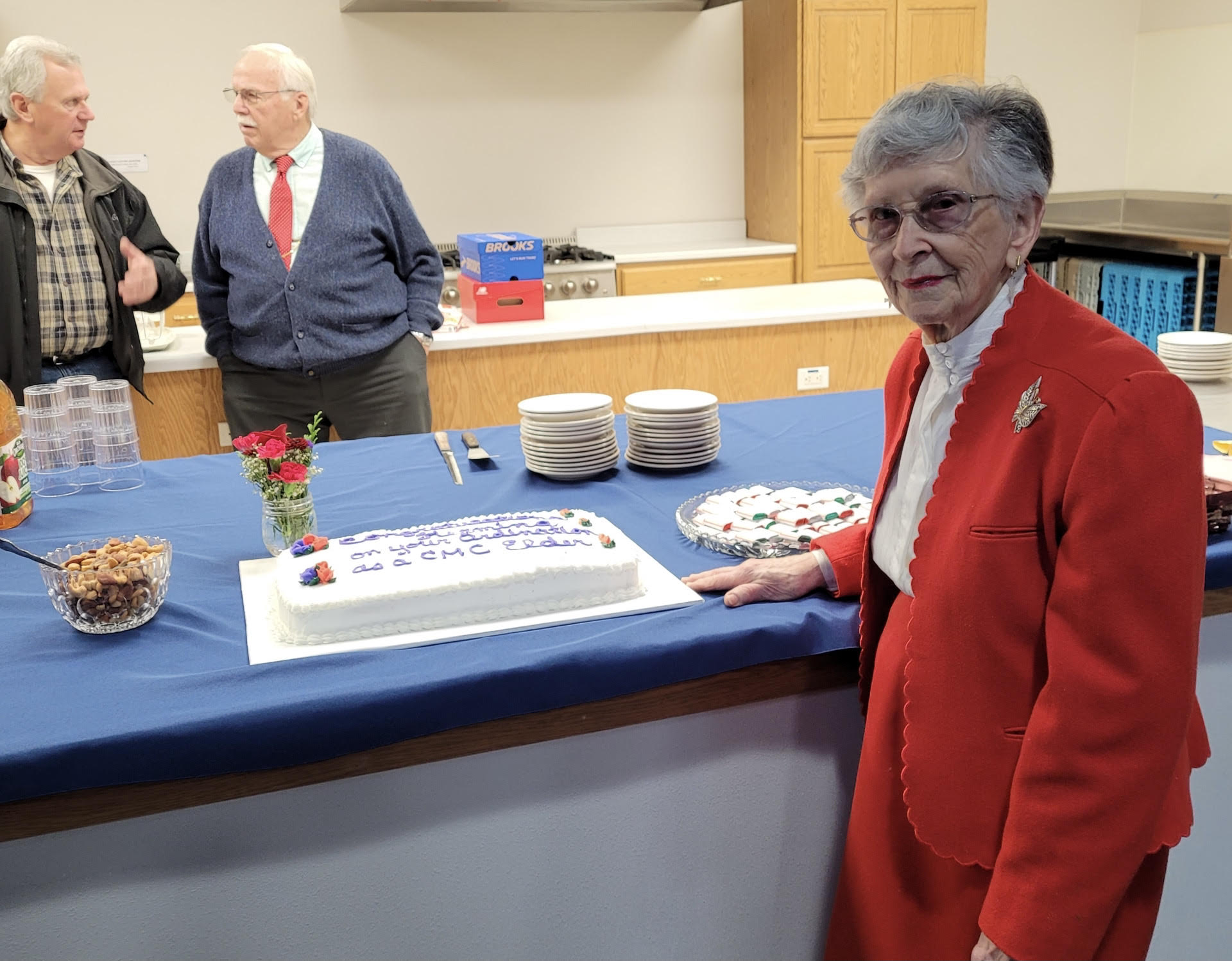 A mature women stands next to a cake while two men talk in the background