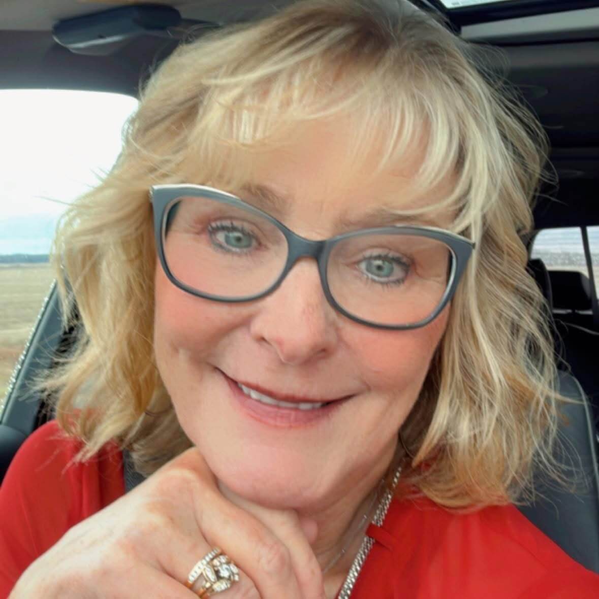 A woman with blonde hair and glasses smiles at the camera while sitting in a car, wearing a red top and a ring on her finger.